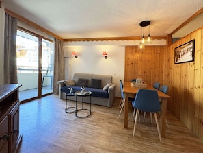 A cozy living room with a sofa, a dining table with chairs, a wooden wall, and a balcony with a view.