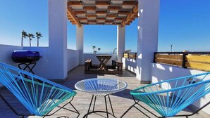 Una terraza en la azotea con dos sillas azules, una mesita redonda y una parrilla.
