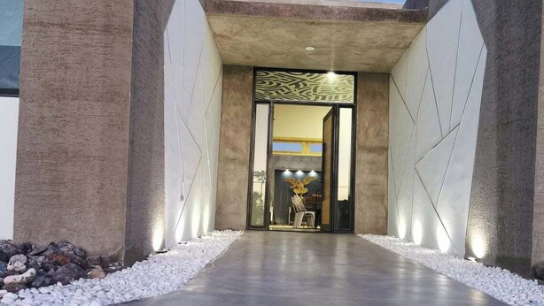 Una entrada moderna con un sendero de concreto, paneles decorativos y un área de descanso con sillas y una mesa.