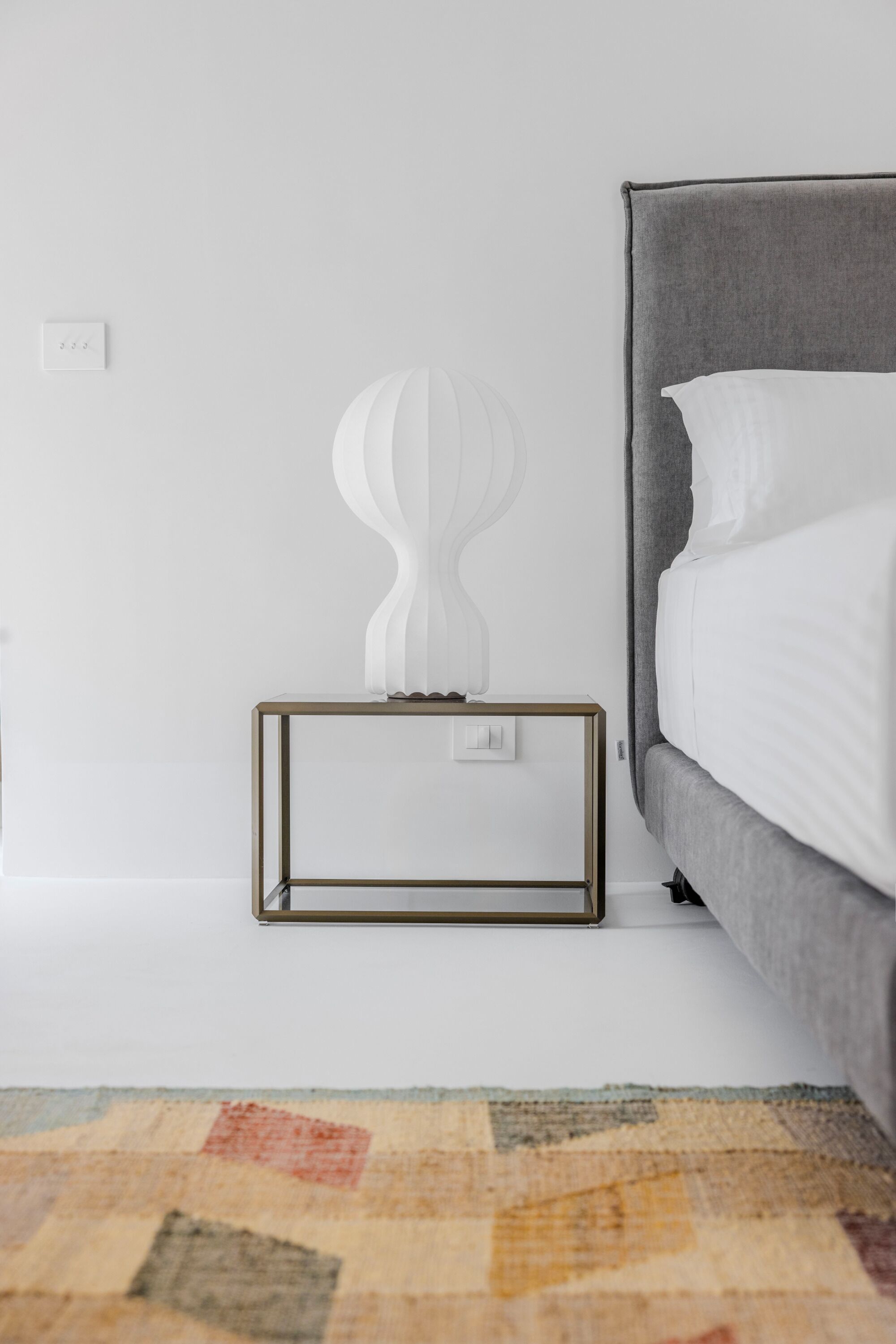 Un dormitorio moderno con una cama tapizada en gris, una mesita de noche blanca con una escultura blanca original y una alfombra estampada en el suelo.