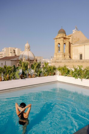 Piscina com uma pessoa na água, cercada por plantas de palmeira e um edifício ao fundo com cúpula e torre sineira.