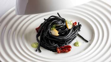 A hand holding a white conical object over a white plate with black noodles, garnished with herbs and a red chili.