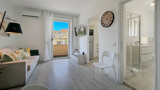 A modern living room with a sofa, a white chair, a flat-screen TV, and a balcony with a view of buildings. - Turchesino Apartment (La Maddalena)