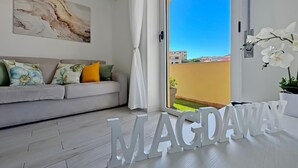 A modern living room with a sofa, decorative pillows, a large window with a view of buildings, and a sign reading "MAGDAWN". - Turchesino Apartment (La Maddalena)