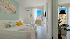 A hotel room with a bed, a bedside table, a mirror, a TV, and a balcony with a view of buildings. - Turchesino Apartment (La Maddalena)
