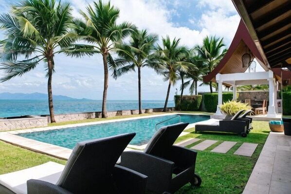Zona junto a la piscina con sillas de descanso, palmeras y vista al océano.