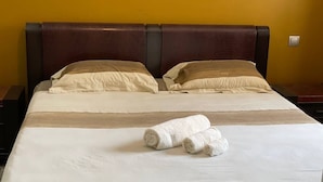 A neatly made bed with two rolled towels, a dark headboard, and a plain yellow wall.