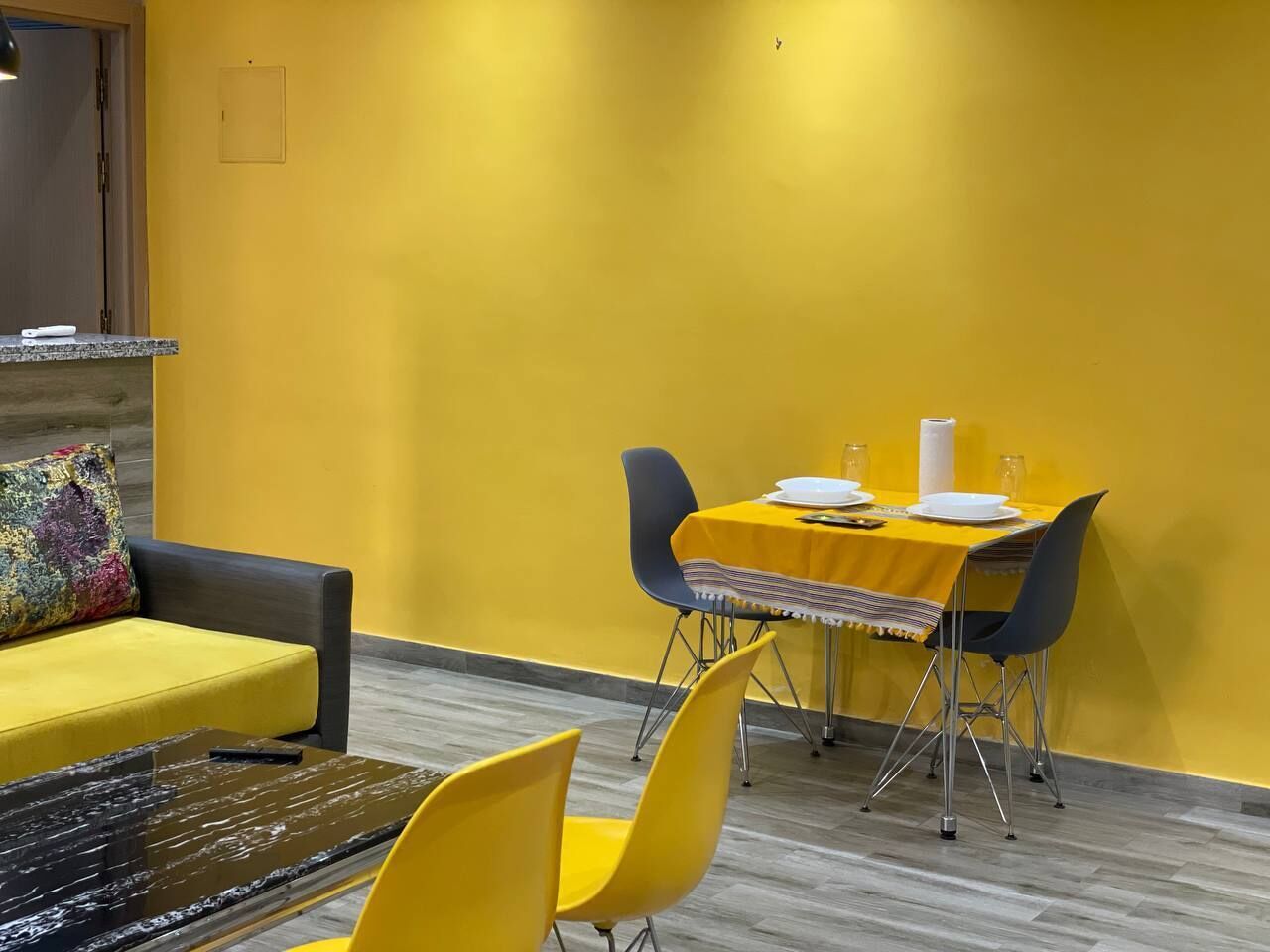 Een eethoek met een gele tafel en stoelen, een bank en een bijzettafeltje met een glazen blad.