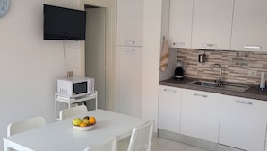 A modern kitchen with white cabinets, a microwave, and a sink. There is a dining table with chairs and a bowl of fruit. - Casa Orchidea (Lu Bagnu)