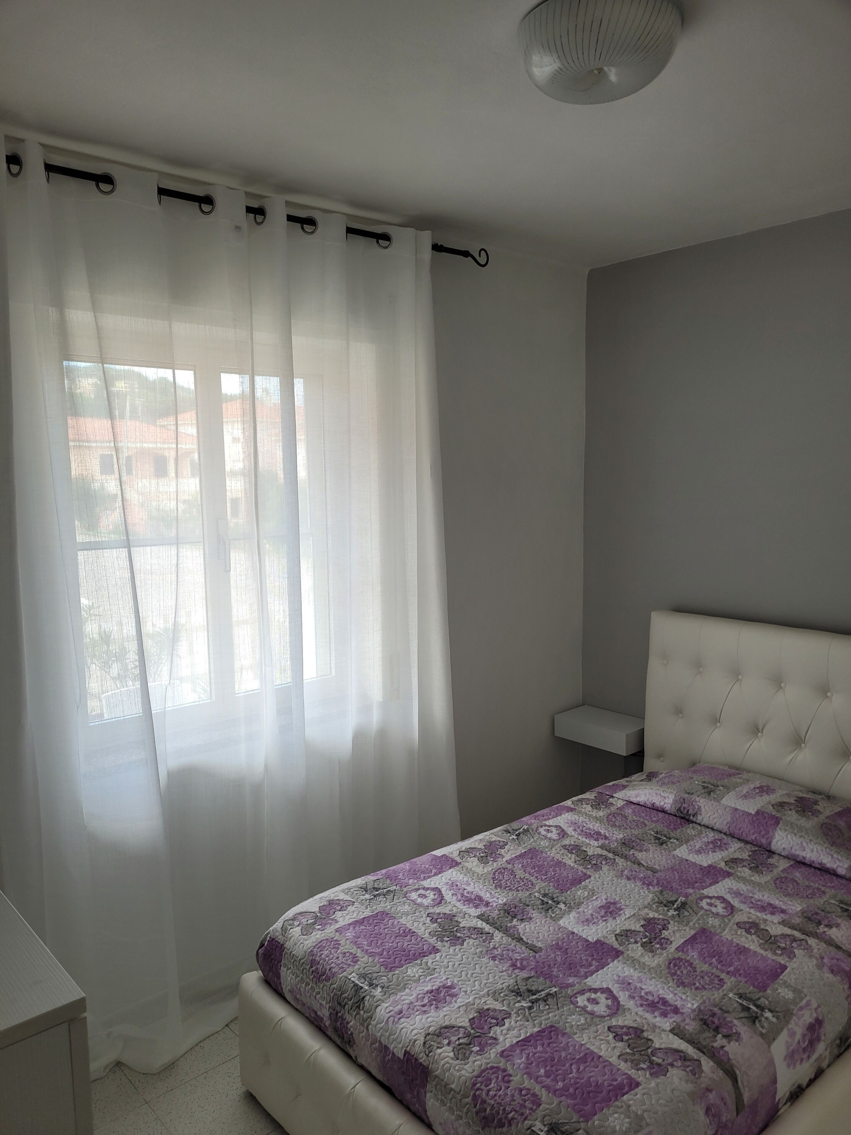 Un dormitorio con cama, una ventana con cortinas traslúcidas, una mesita de noche y una lámpara en el techo.