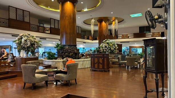 Een ruime lobby met hoge houten pilaren, comfortabele zithoeken en decoratieve bloemarrangementen.