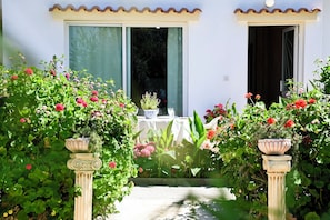 A building with a white facade, large windows, and a door. There are potted plants with flowers in front of the entrance. - Room-kritos Apartments-one Bedroom (Argaka)