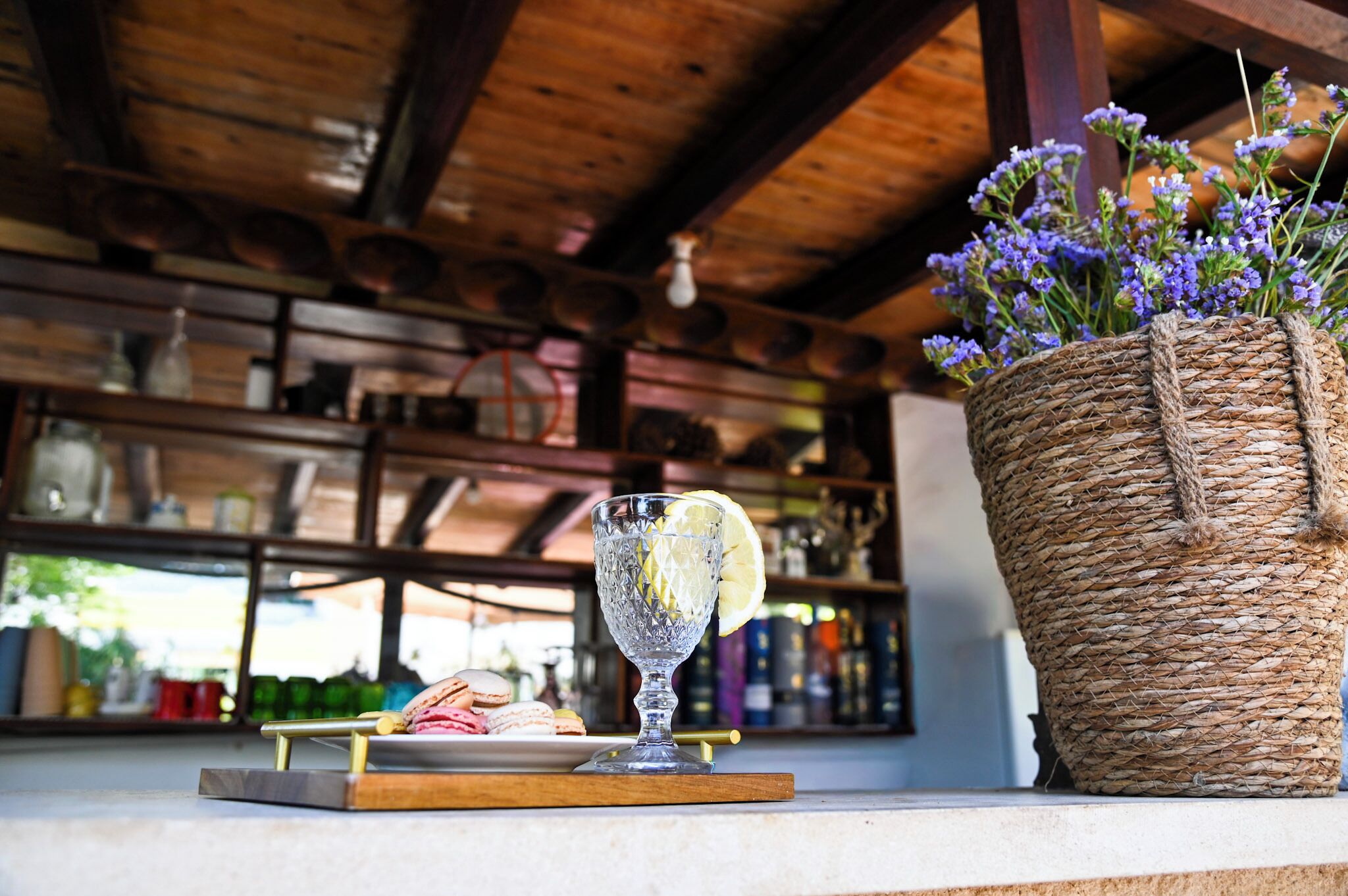 Un vaso con una rodaja de limón sobre una bandeja, una cesta tejida con flores y estantes con diversos objetos.