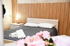 A neatly made bed with white pillows and a folded towel, a wooden slatted headboard, a nightstand with a lamp, and a vase with flowers. - Room-kritos Apartments-one Bedroom (Argaka)