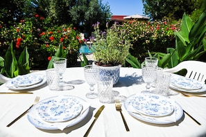 An outdoor dining table set with blue and white plates, gold cutlery, and crystal glasses, surrounded by lush greenery and flowers. - Room-kritos Apartments-one Bedroom (Argaka)