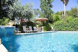 A swimming pool with lounge chairs and an umbrella, surrounded by greenery and trees. - Room-kritos Apartments-one Bedroom (Argaka)