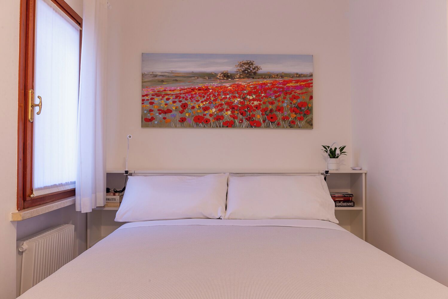 Un dormitorio con cama, ropa de cama blanca, un cuadro de un campo de flores en la pared, una planta pequeña en una mesita de noche y un radiador.