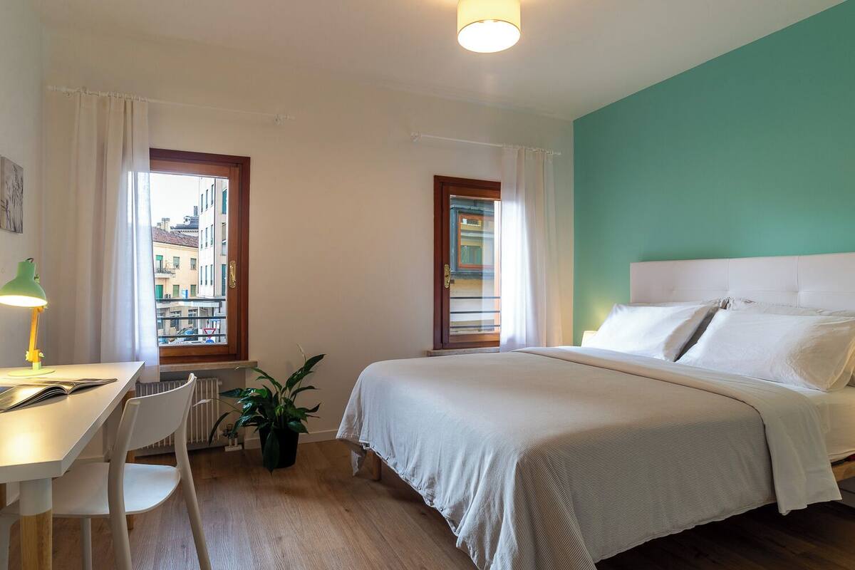 A hotel room with a bed, a desk with a lamp, a chair, and a potted plant.