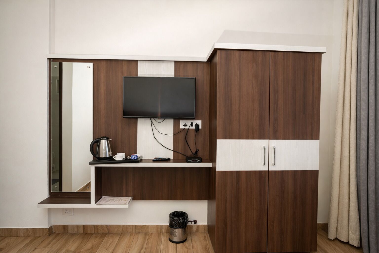 A hotel room with a TV mounted on a wooden panel, a small shelf with a kettle, a trash bin, and a wardrobe with two doors.