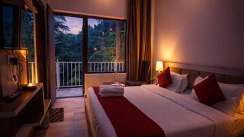 A hotel room with a large bed, a nightstand, a lamp, a chair, and a balcony with a view of trees.