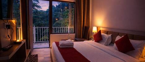 A hotel room with a large bed, a nightstand, a lamp, a chair, and a balcony with a view of trees.