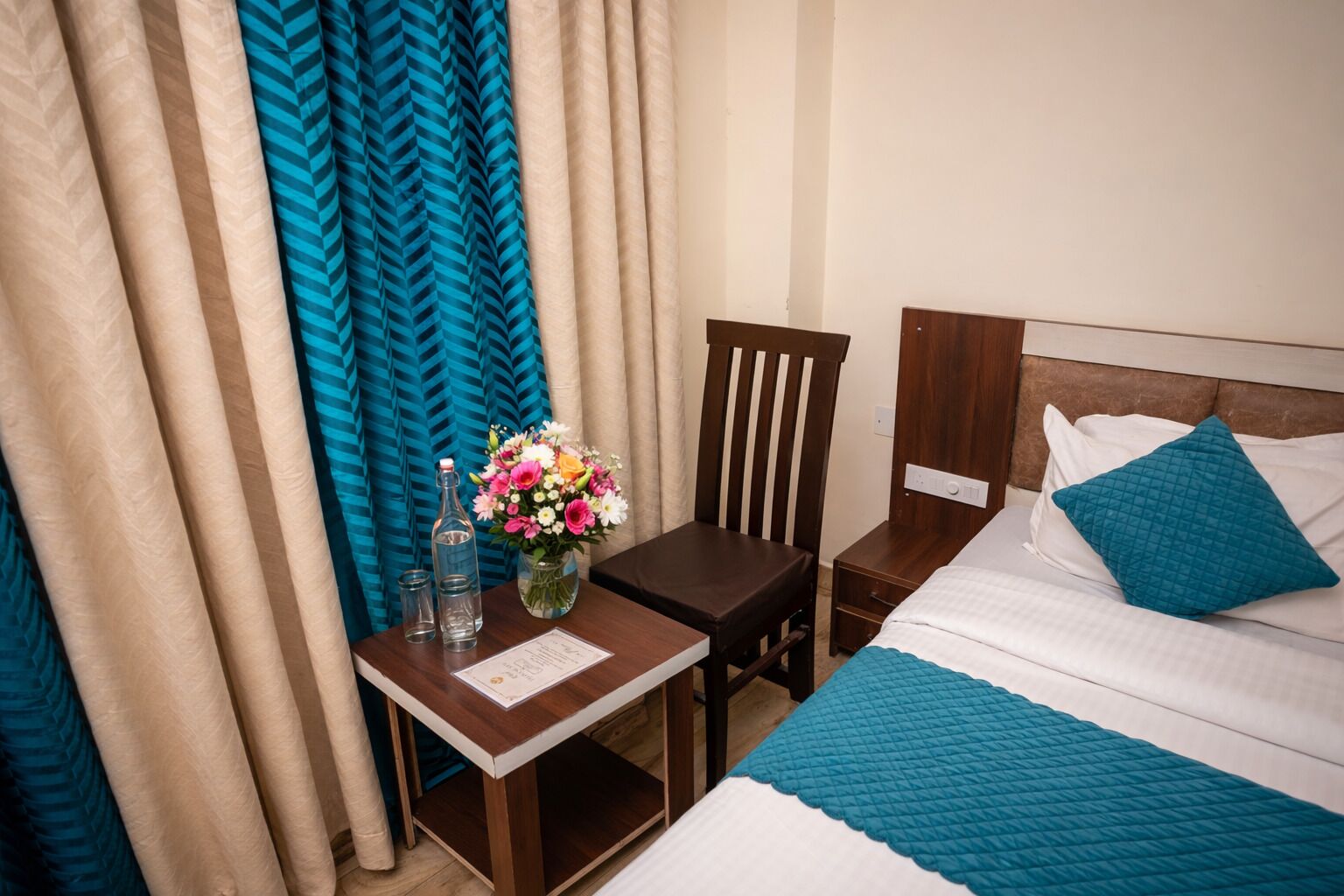 Una habitación de hotel con una cama, una mesita de noche de madera, una mesa con un florero y una silla.