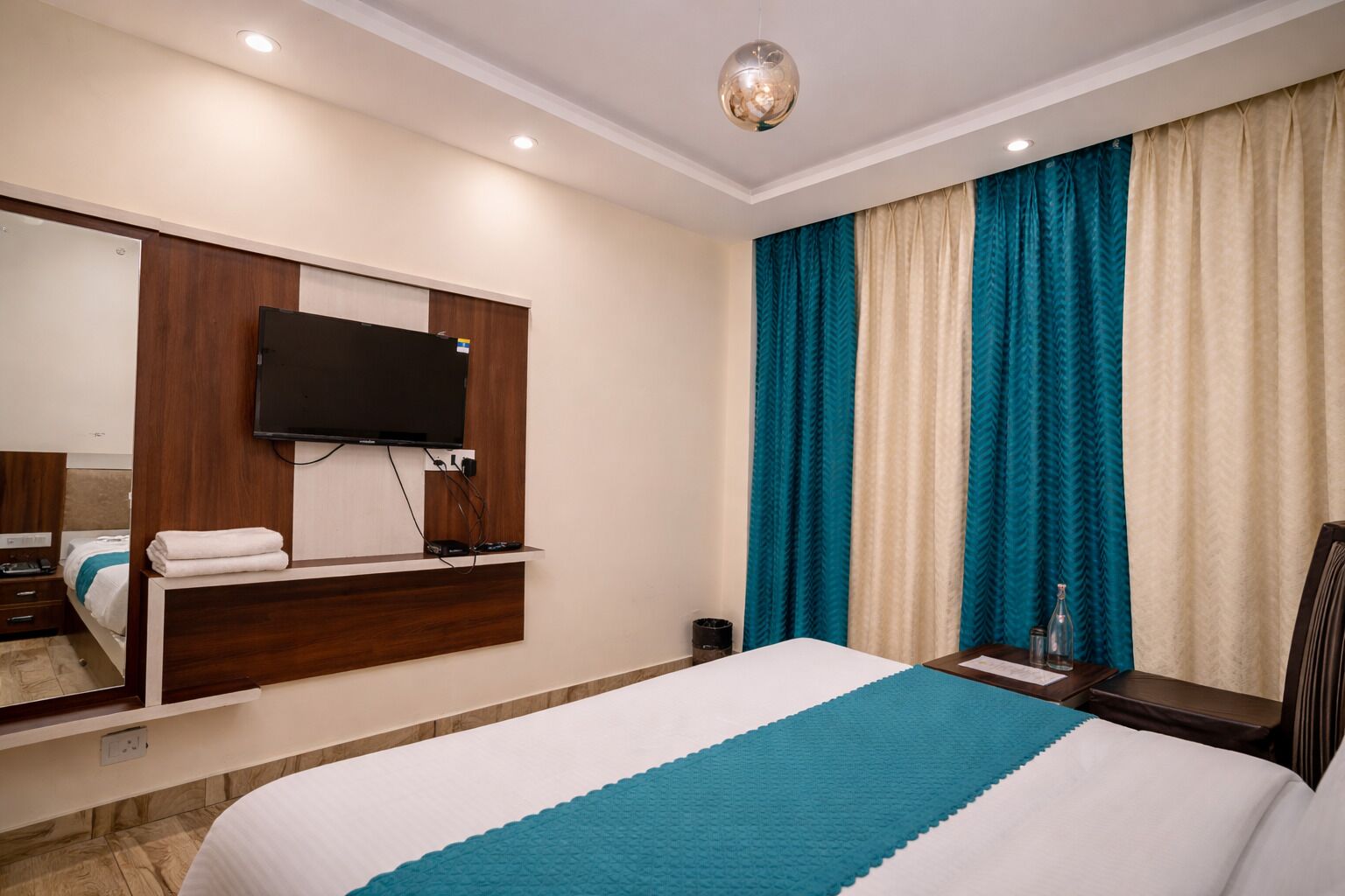 Una habitación de hotel con una cama, un televisor montado en un panel de madera, una mesita de noche con una lámpara y una ventana con cortinas.