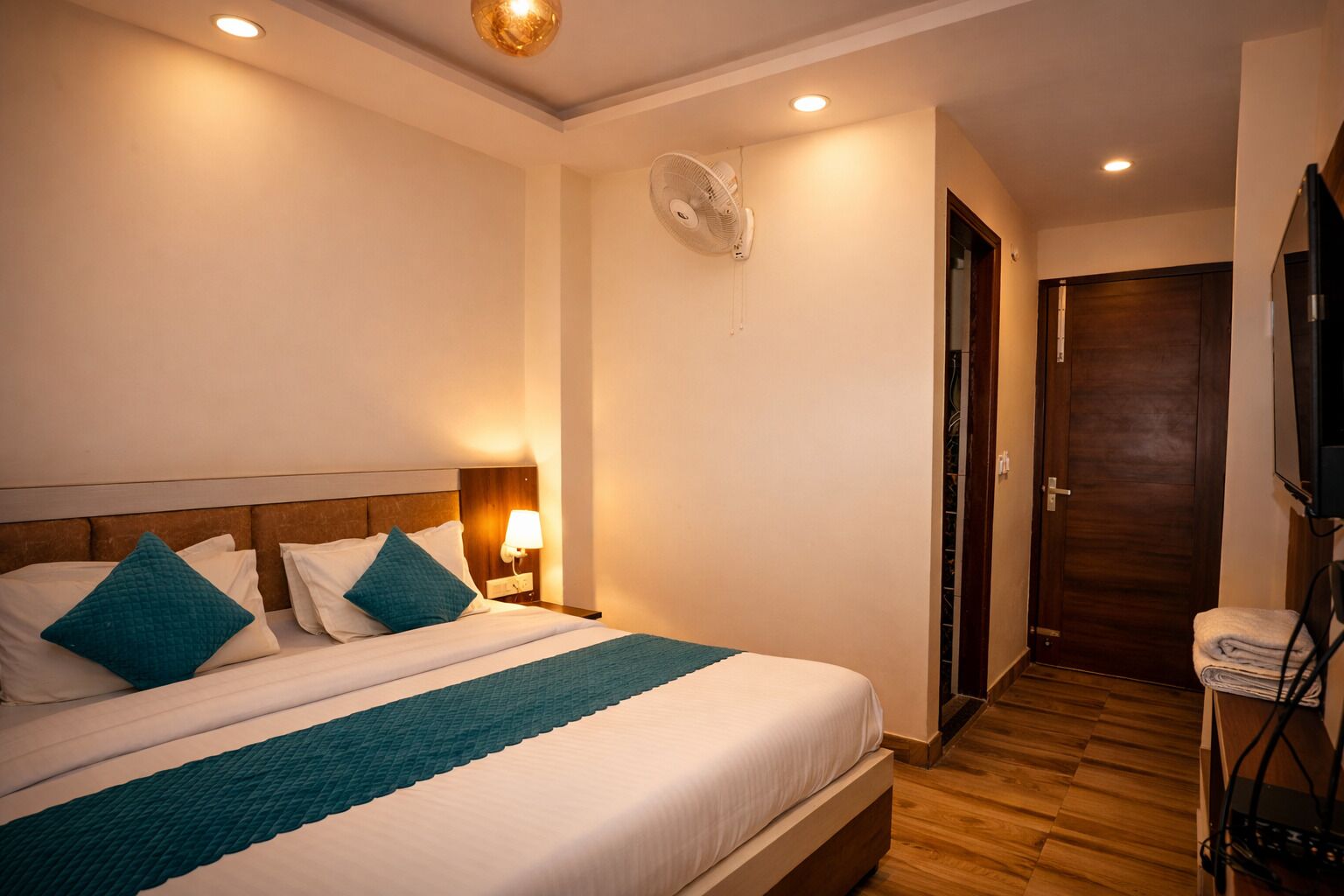 A hotel room with a bed, bedside lamp, ceiling fan, wooden floor, and a door.