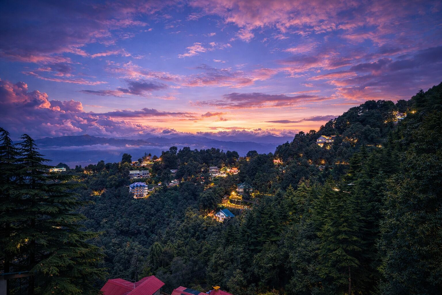 A mountainous landscape with scattered buildings, dense forest, and a sunset sky.