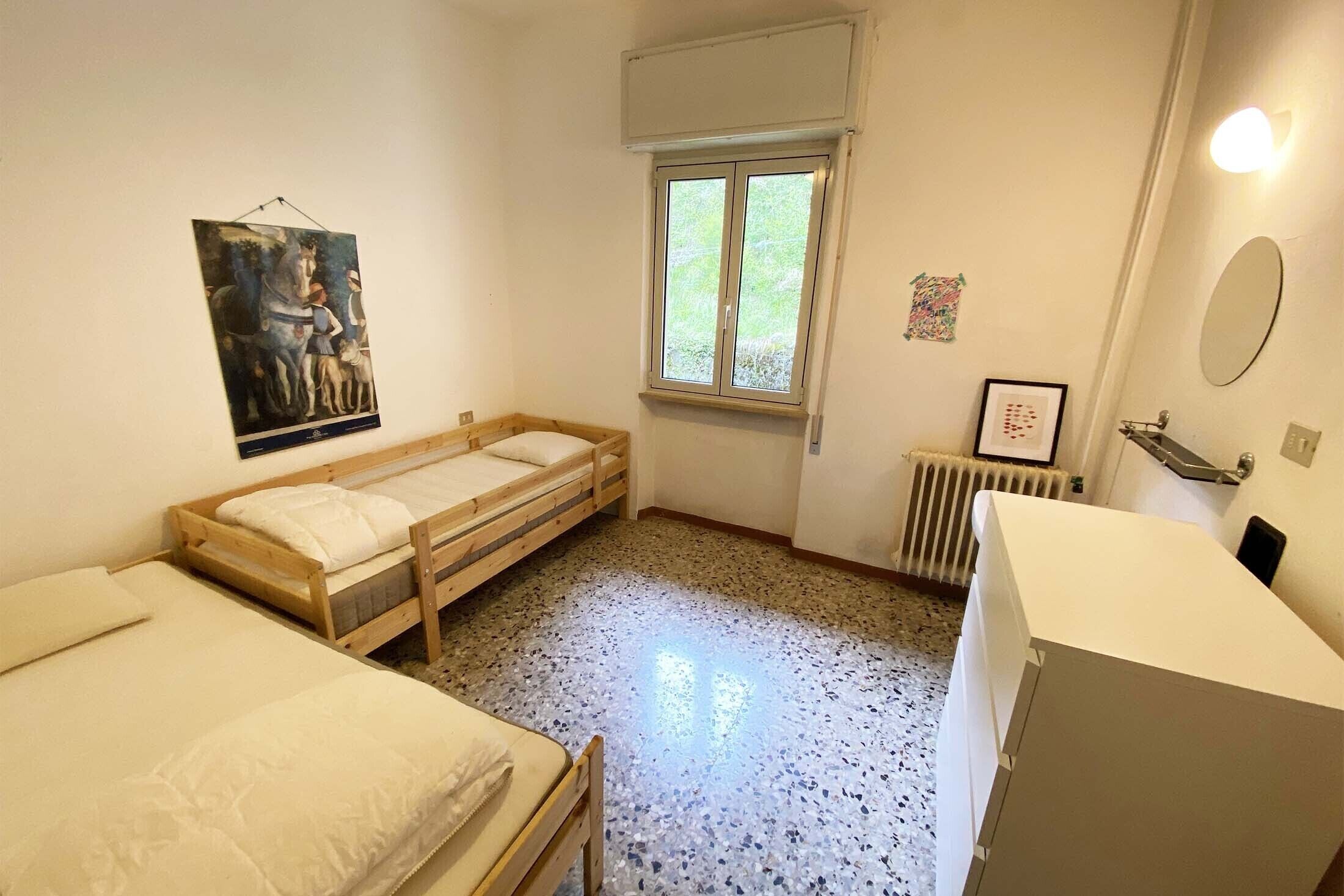 Una stanza piccola e pulita con due letti di legno, una finestra, un termosifone e uno specchio al muro.