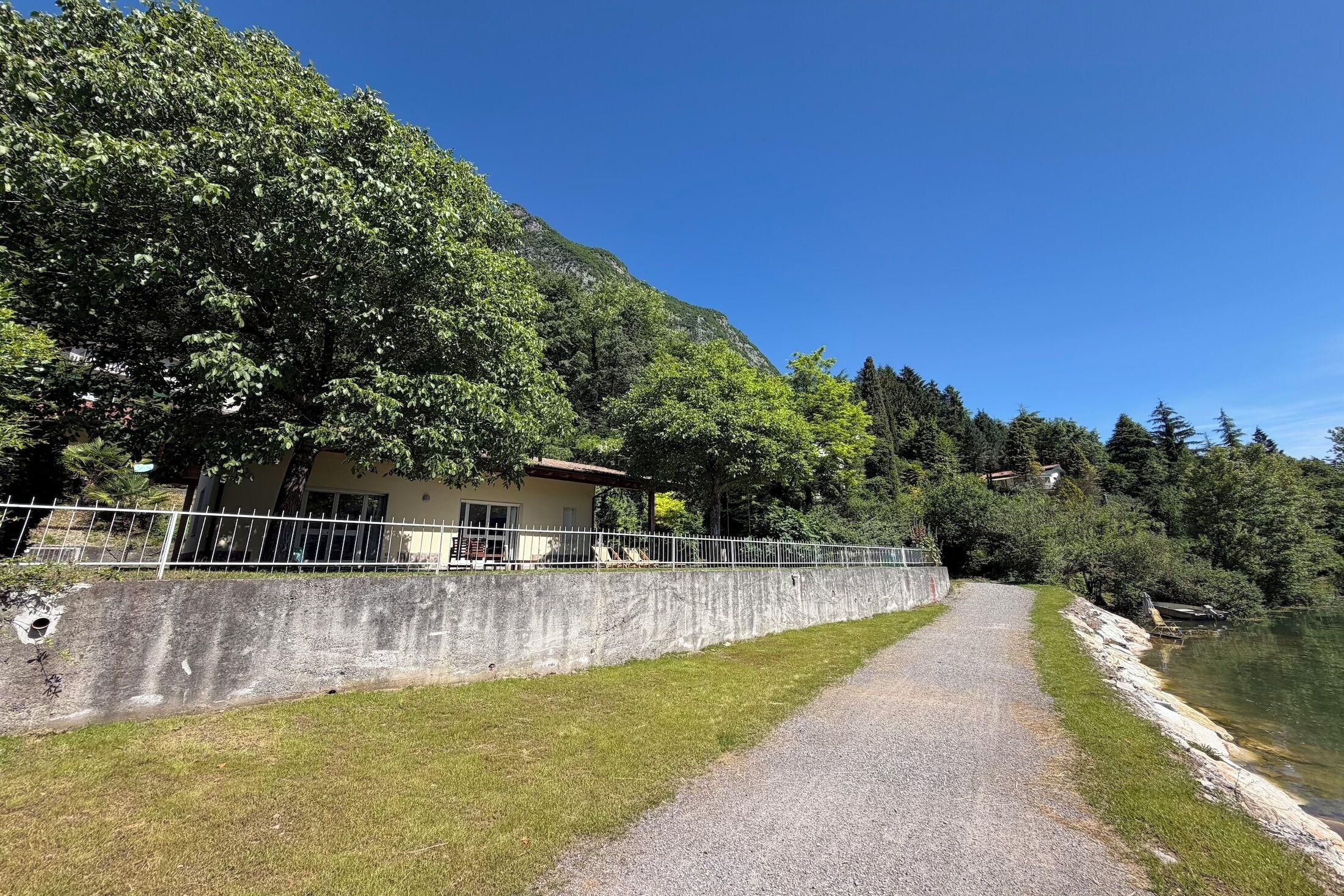 Un sendero de grava que conduce a un edificio con un muro de concreto y una cerca de metal, rodeado de árboles y una masa de agua.