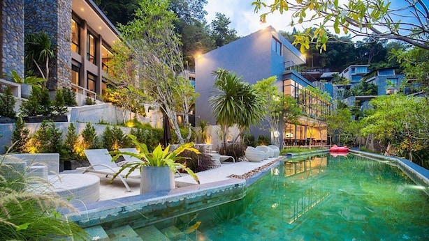 A modern pool area with clear water, surrounded by lush greenery and stone steps leading to a building with large windows. - Design Duplex P10 in Natural Park Resort (Kamala)