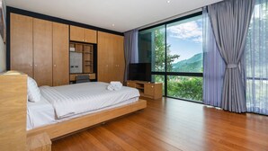 A modern bedroom with a wooden bed, built-in wardrobe, a TV, and a large window with a view of greenery. - Design Duplex P10 in Natural Park Resort (Kamala)