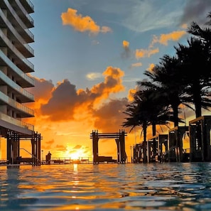 A waterfront hotel with a pool reflecting the sunset, a pedestrian bridge, and palm trees. - 39th Floor Oceanfront Penthouse Hollywood Beach · Resort Pool · Stunning Views (Hollywood)