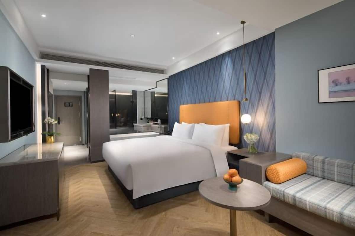 A modern hotel room with a large bed, a sofa, a small table with fruit, a TV, and a bathroom visible through an open door.