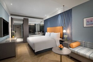 Une chambre d’hôtel moderne avec un grand lit, un canapé, une petite table avec des fruits, une télévision et une salle de bain visible à travers une porte ouverte.
