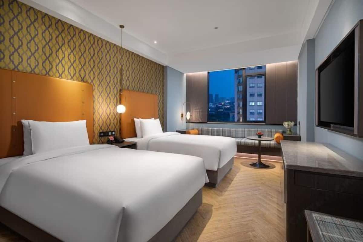 A hotel room with two beds, a TV, a small table, and a view of the city at night.