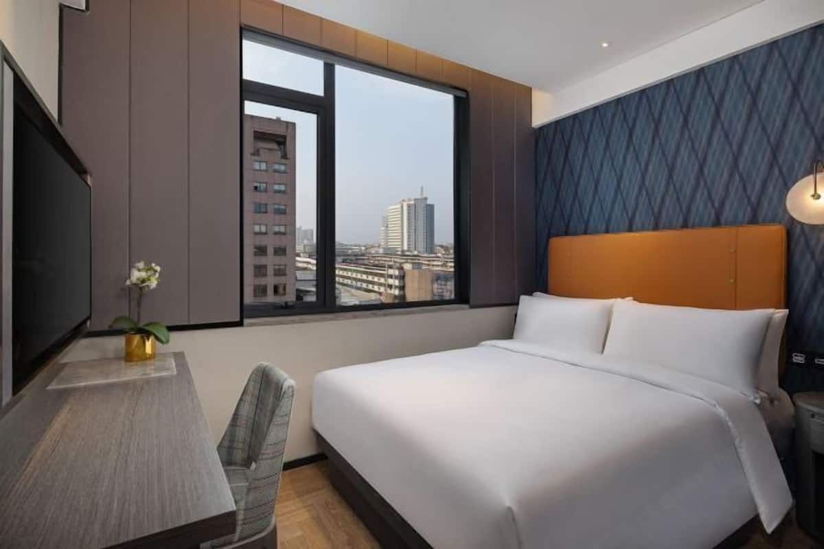 A hotel room with a large bed, a desk with a chair, a television, and a window with a city view.