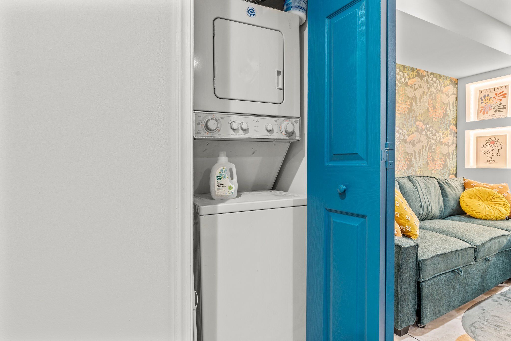 A laundry room with a washing machine, a blue door, and a grey sofa with yellow pillows.