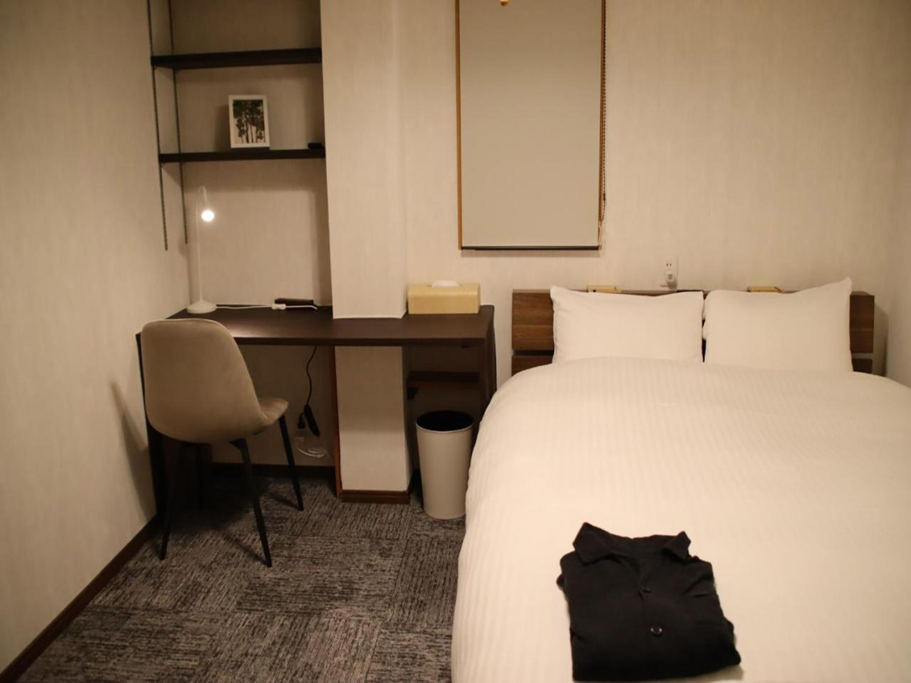 A hotel room with a bed, a desk, a chair, a trash bin, and a shelf with a picture.