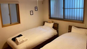 Ein Hotelzimmer mit zwei Einzelbetten, einem großen Fenster mit Jalousien, einer hängenden Pendelleuchte und einem kleinen Nachttisch mit Lampe.