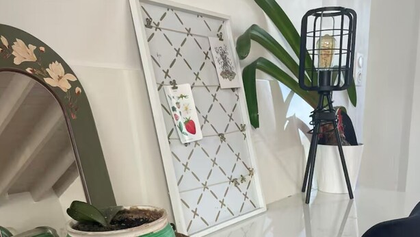 A white table with a woven basket containing a green plant, a framed picture with a geometric pattern, and a mirror with floral design. - Charming house in the heart of the village (Bazouges-la-Pérouse)