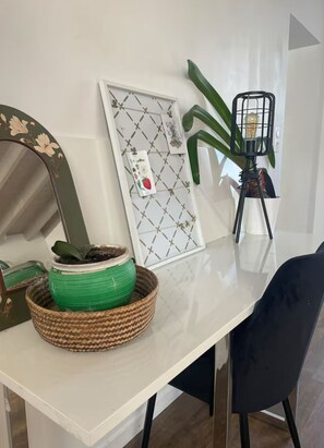 A white table with a woven basket containing a green plant, a framed picture with a geometric pattern, and a mirror with floral design.