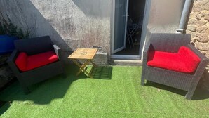 An outdoor patio with two wicker chairs with red cushions, a small wooden table, and a door leading to a covered area. - Charming house in the heart of the village (Bazouges-la-Pérouse)