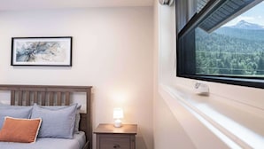 A bedroom with a bed, a nightstand with a lamp, a framed abstract artwork, and a window with a mountain view. - The Cedar Suite - Secluded Mountain Retreat with Forest Views and Hot Tub (Revelstoke)