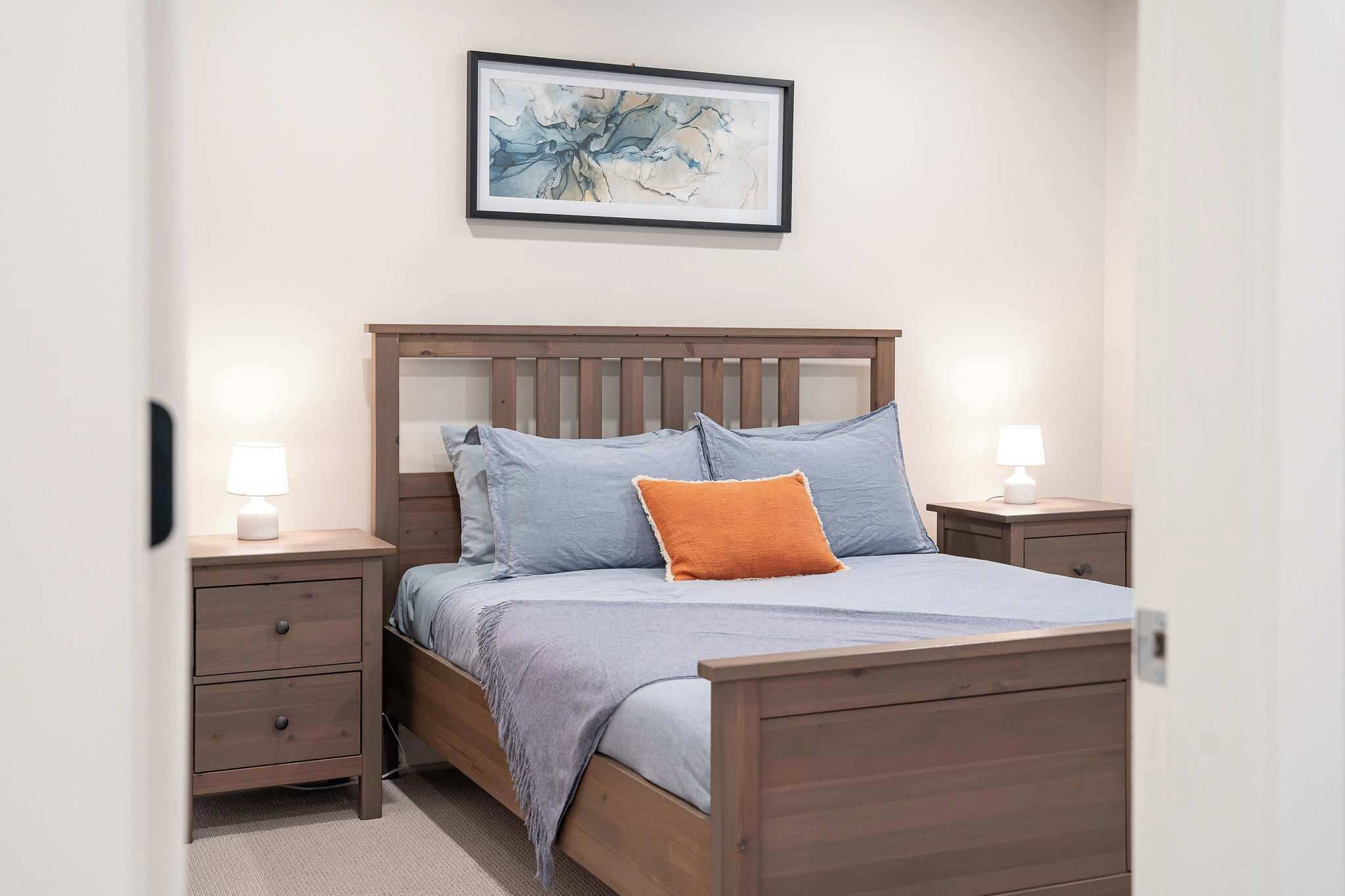 Een slaapkamer met een houten bed, twee nachtkastjes met lampen, een schilderij met abstracte kunst aan de muur, en een vloerbedekking.