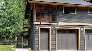 A two-story building with a balcony, large garage doors, and a wooden deck. - The Cedar Suite - Secluded Mountain Retreat with Forest Views and Hot Tub (Revelstoke)