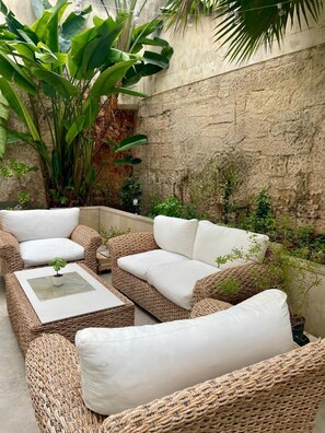 Eine Sitzgruppe im Freien aus Korbmöbeln mit einem weißen, gepolsterten Sofa und einem kleinen Tisch, umgeben von üppigem Grün und einer Steinmauer.