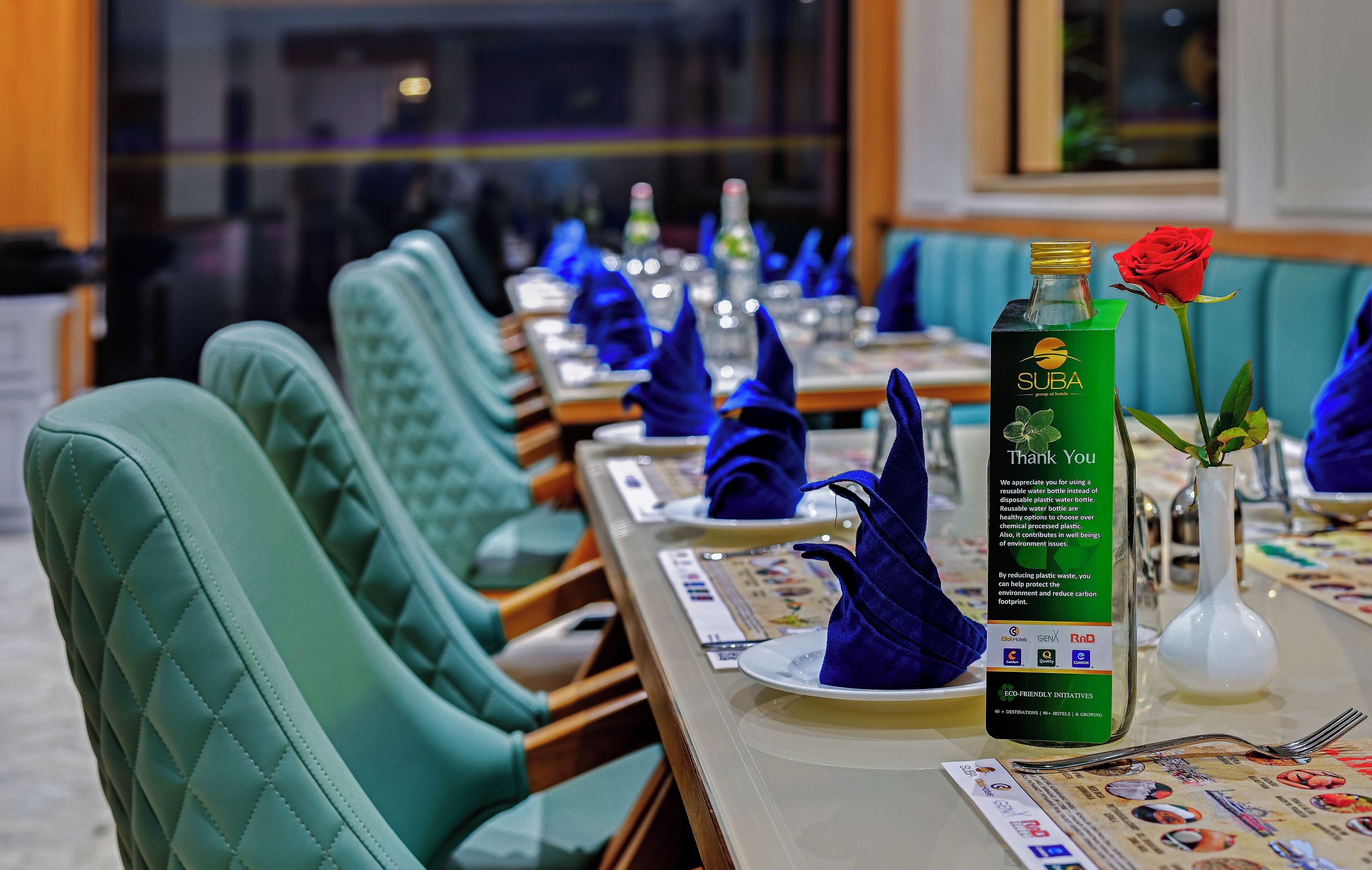 Un coin repas avec des chaises recouvertes d’un tissu turquoise, des tables soigneusement dressées avec des serviettes bleues, une bouteille verte ornée d’une rose et un menu.
