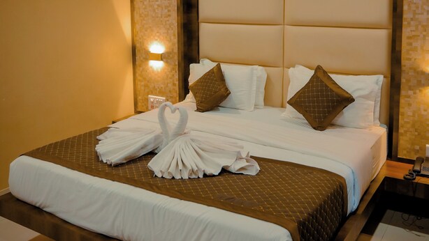 A hotel room with a large bed, a ceiling fan, an air conditioning unit, and a nightstand with a lamp. - Hotel Royal Celebration (Kawardha)